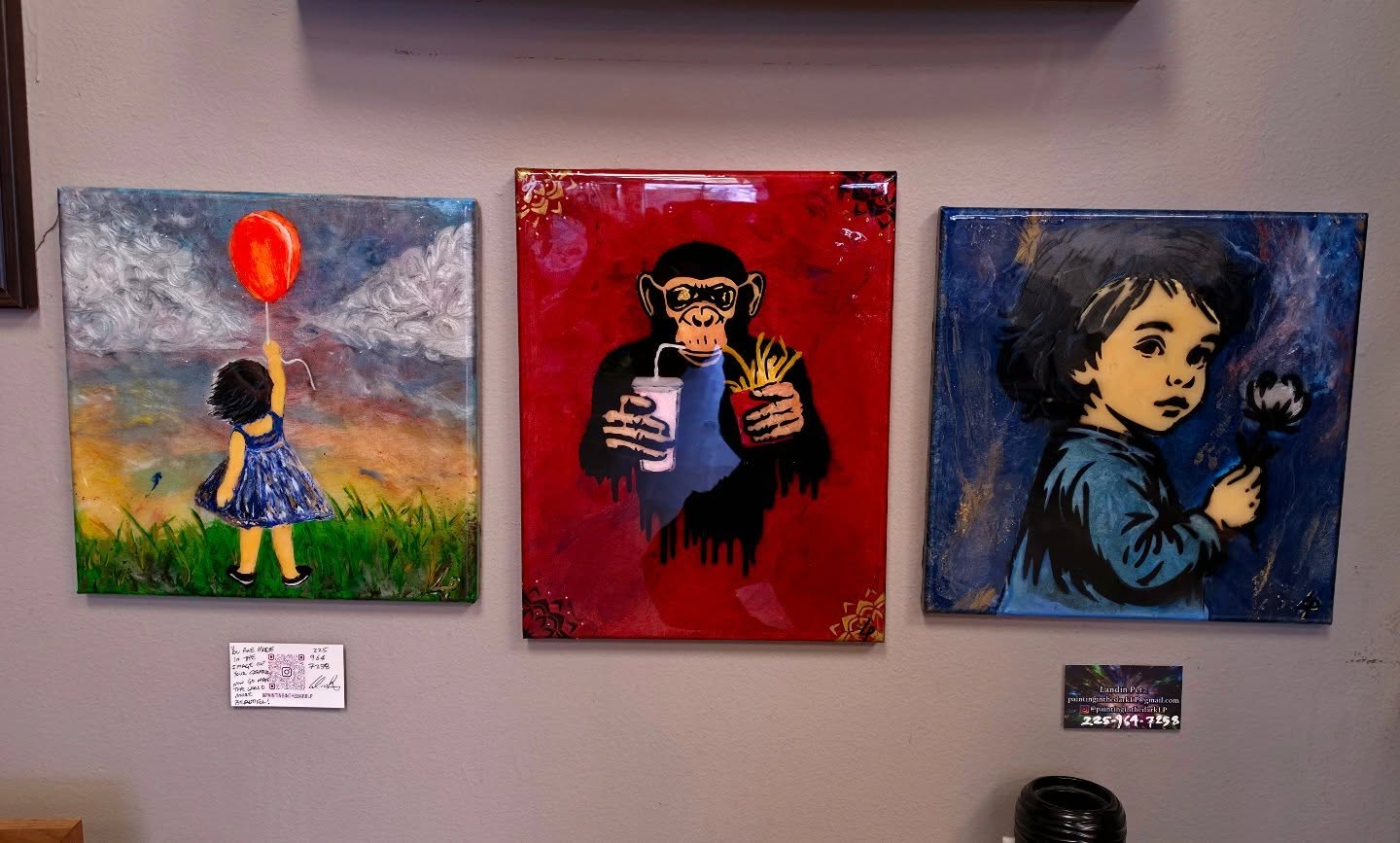 Three original paintings displayed at The Bearded Barber, including a child with a balloon, a chimpanzee, and a child holding a flower